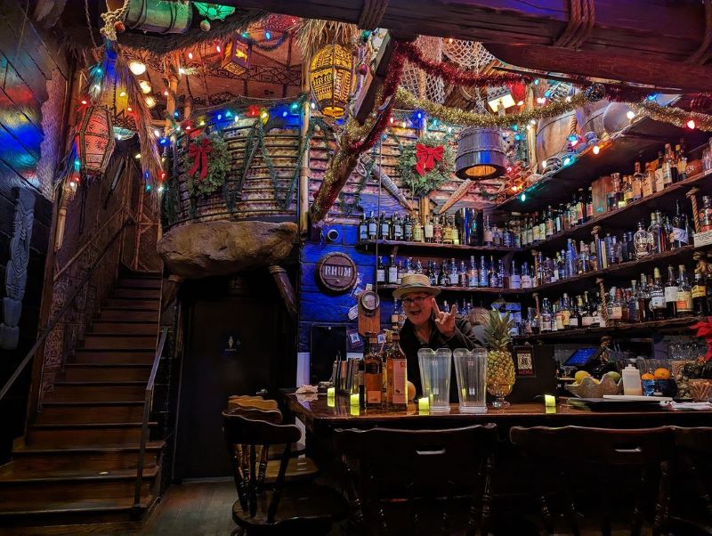A bartender gives a shocka sign from behind the bar at Smuggler's Cove, whose walls are lined with liquor bottles, colorful lanterns, and tiki decor. 