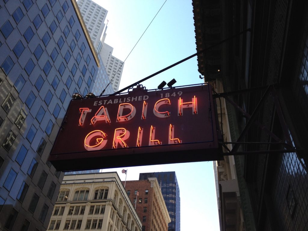 A sign that reads Tadich Grill with the SF skyline in the background