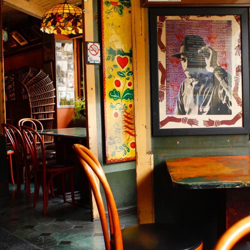 Wooden tables and chairs at Vesuvio Bar with colorful art on the walls and a stained glass lamp.