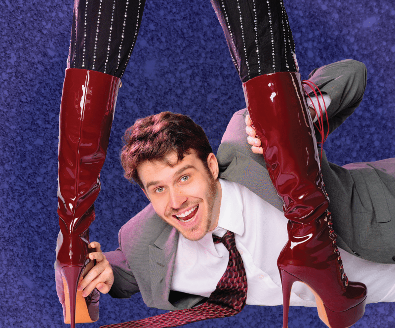 An actor portraying Charlie lays on the ground peeking through the legs of a person wearing knee-high red stilettos. 