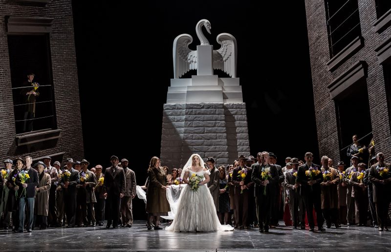A bride dressed in white stands center-stage in front of a crowd of people in dark clothing and a statue of a swan.