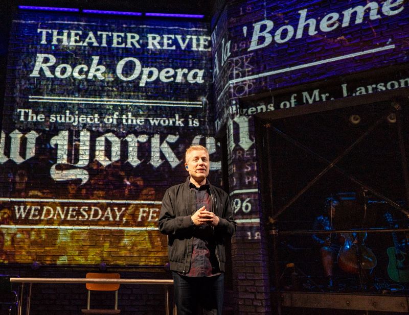 Anthony Rapp performs in front of projected press headlines. 