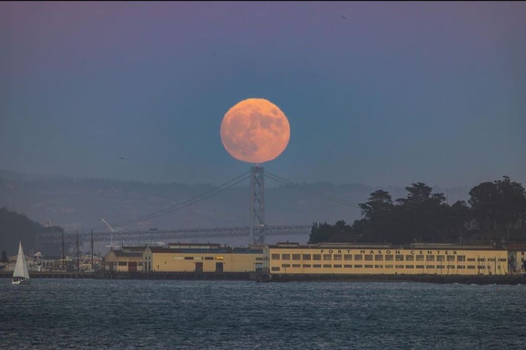 A large orange-tinted moon rises over the Bay Bridge.